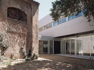 A courtyard blending old and new architecture, with a historic building on the left and a modern glass structure on the right, surrounded by greenery and shaded trees.