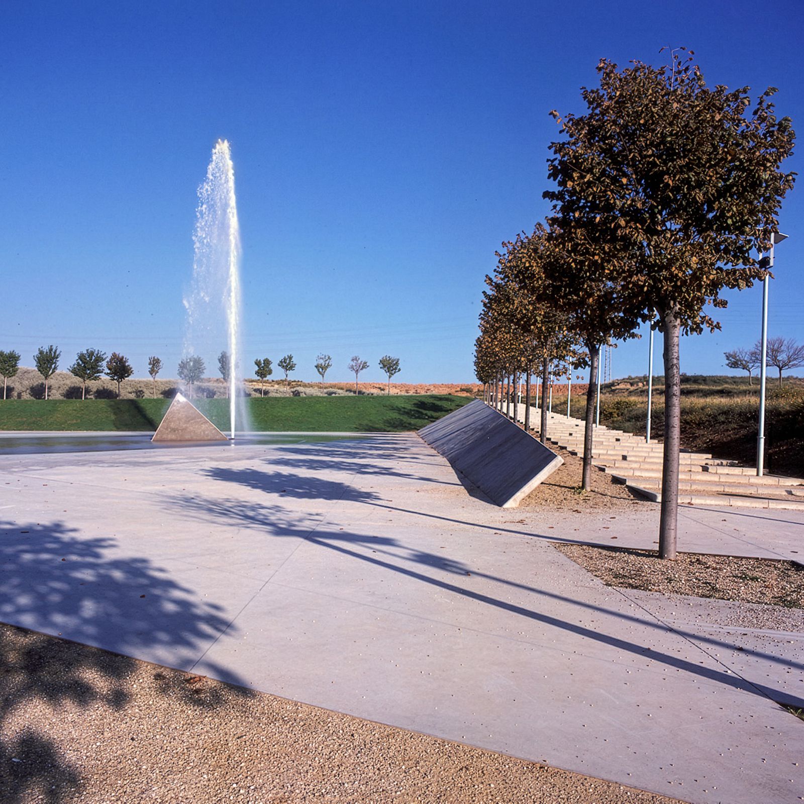 Una línea de árboles a lo largo de un camino con escalones suaves junto a una gran fuente de agua con chorro en un área de parque abierto.