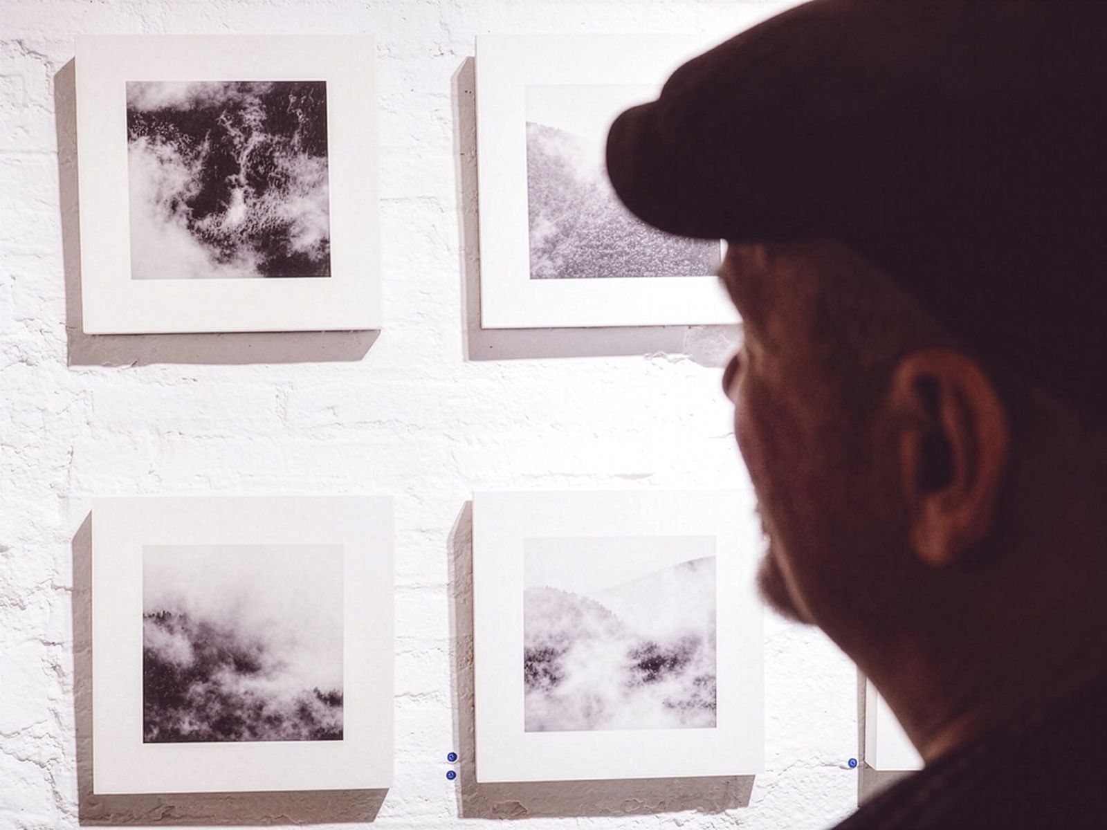 Una persona mirando una serie de fotografías cuadradas en blanco y negro de nubes y niebla en una pared de ladrillo blanco.