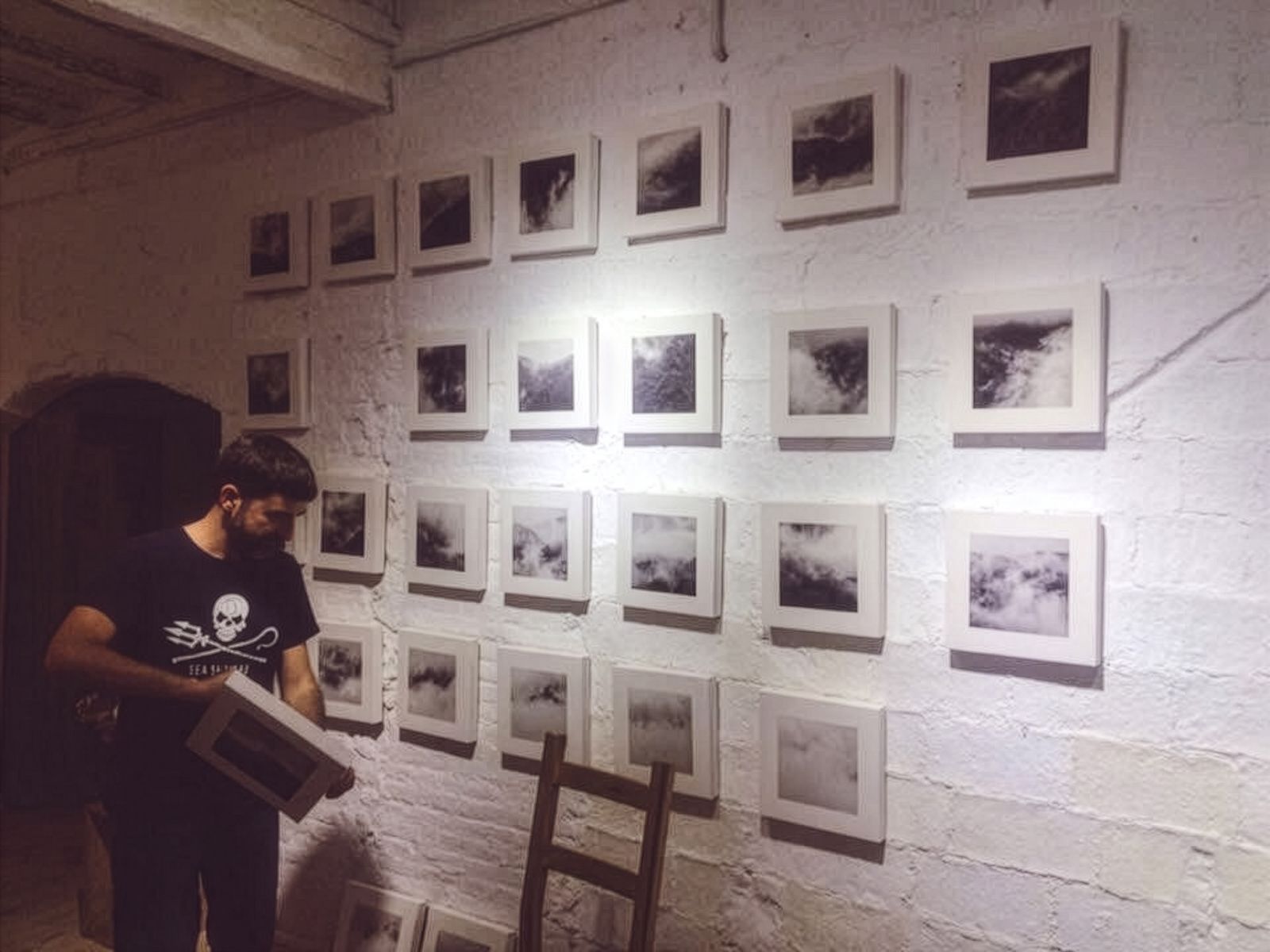 Un hombre colocando fotografías en blanco y negro en una pared de ladrillo blanco, con varias ya expuestas en un patrón de cuadrícula, en un ambiente de galería.