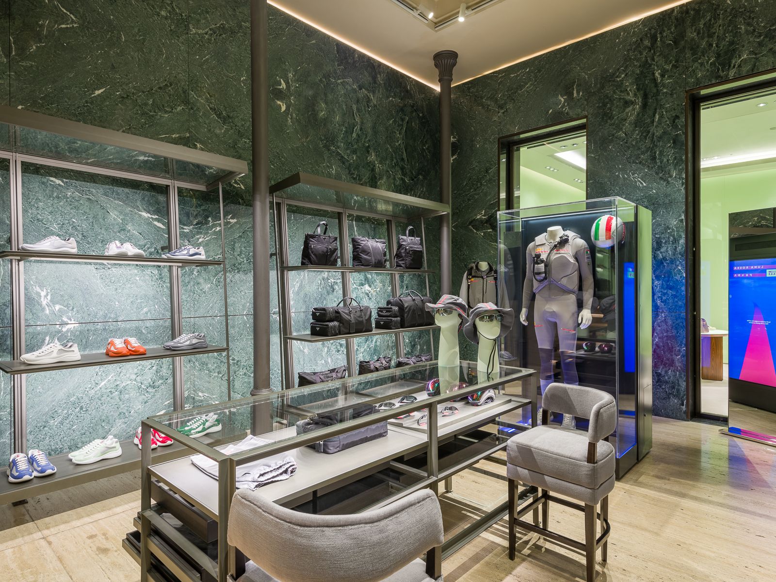 High-end boutique interior with green marble walls, sleek metal shelving showcasing sneakers and bags, and a glass display case featuring a mannequin in a performance outfit with a racing helmet. A central glass table holds accessories, with plush seating nearby.