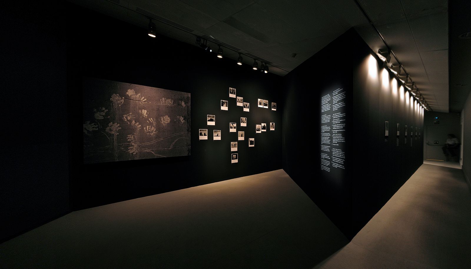 Wide-angle photograph of a dimly lit art exhibition space with matte black walls, featuring a large artwork showing white handprints, a cluster of small framed photographs, and an adjacent illuminated corridor.