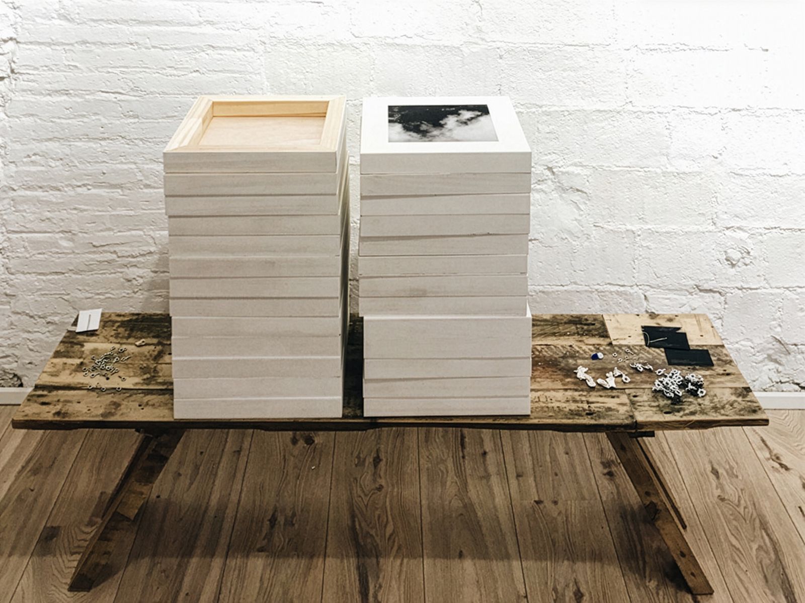 Dos pilas de fotografías enmarcadas en blanco sobre una mesa de madera, junto a pequeños ganchos metálicos y otros materiales de colgado, en una galería con paredes de ladrillo blanco.
