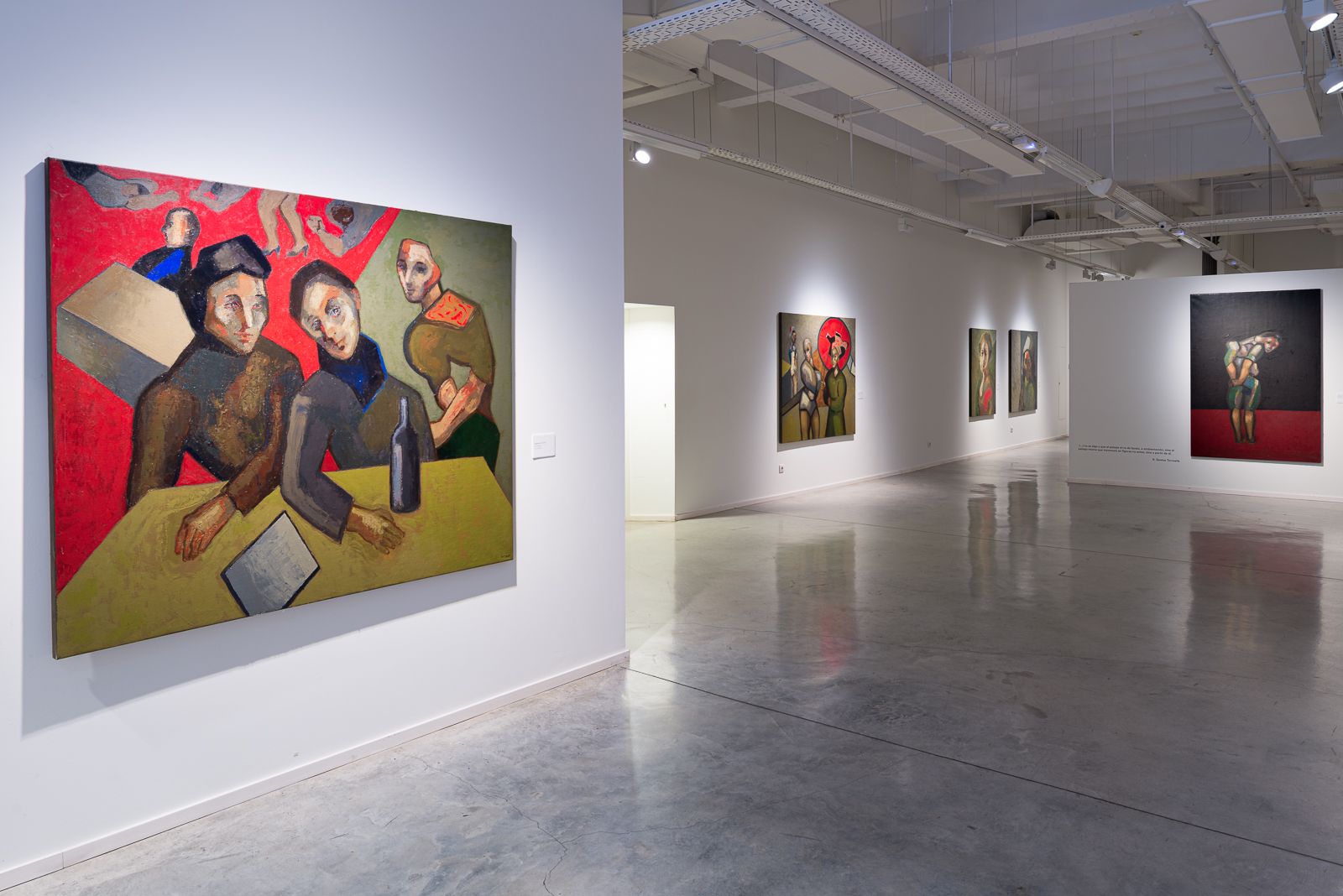 Gran espacio de exposición con pinturas coloridas de Guerrero Medina, incluyendo una pieza destacada que representa tres figuras en una mesa.