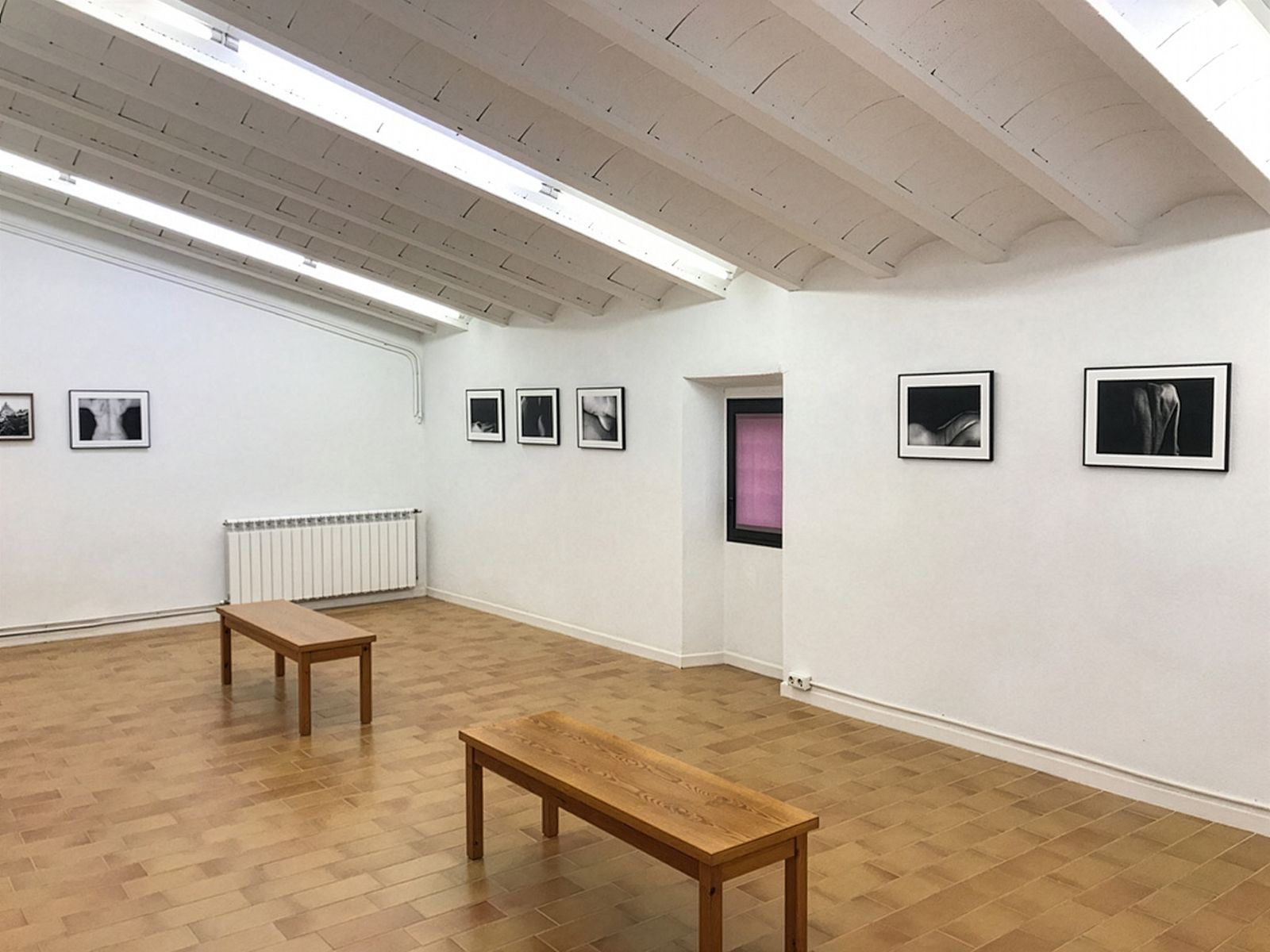 A minimalist gallery room with black-and-white photographs on white walls, two wooden benches in the center and a small window on one wall.