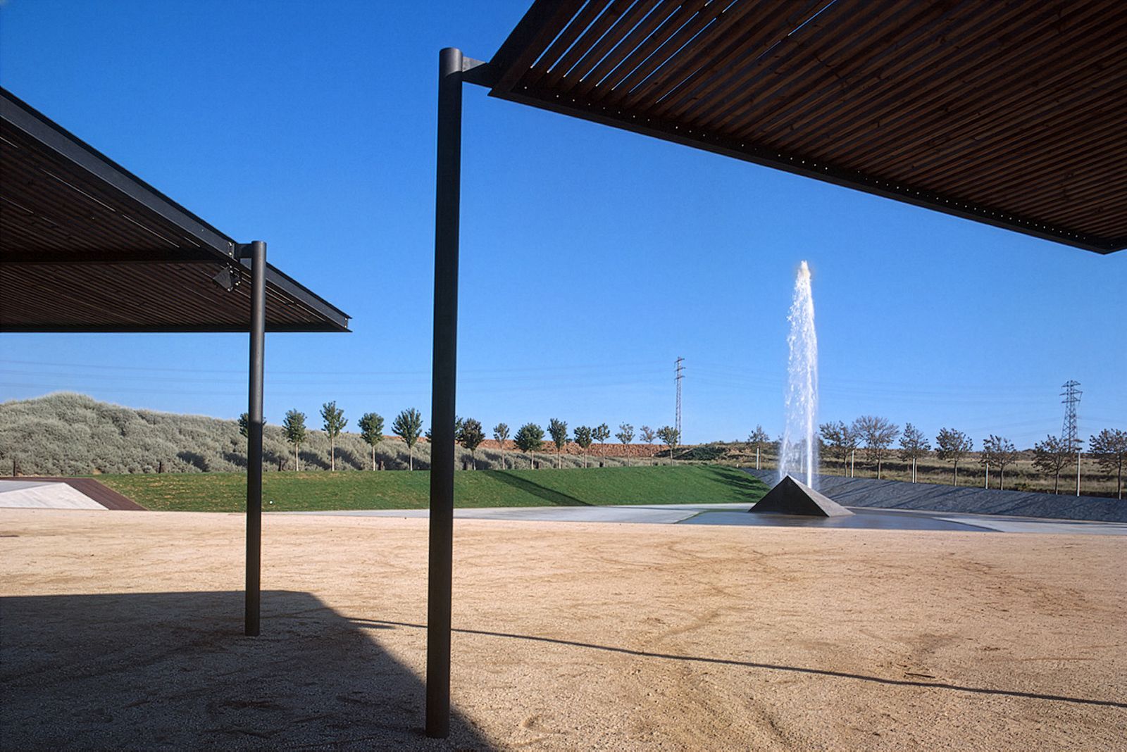 Espacio al aire libre con dos modernas estructuras de sombra de acero y madera y una gran fuente de agua con chorro central contra un cielo azul despejado.