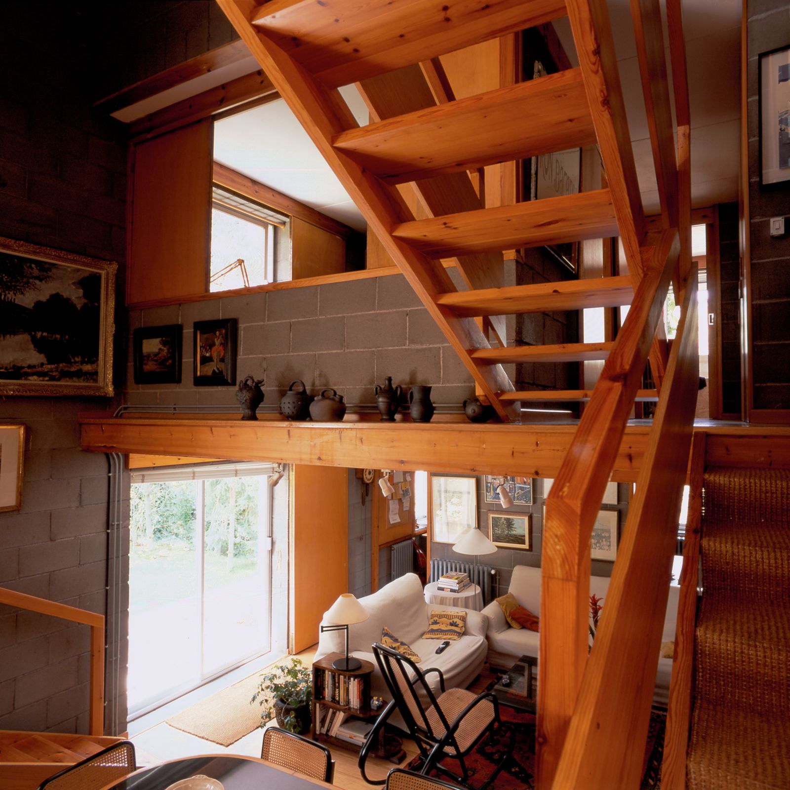 Interior view showing a wooden staircase, a cozy living room with white sofas, a rocking chair, a rug, and decorative items on shelves and walls. The room has concrete block walls and a large sliding door letting in natural light.