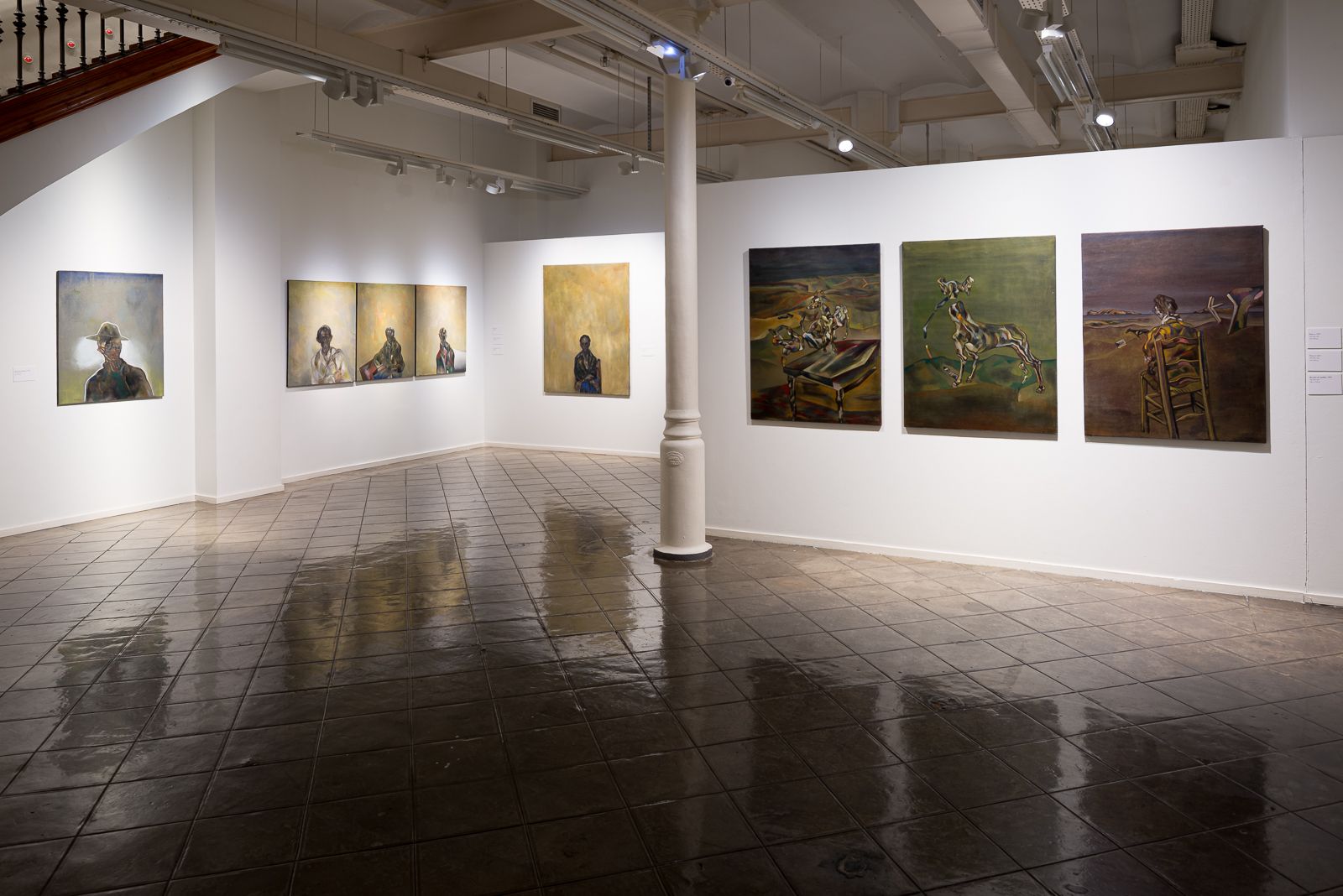 Un rincón de galería con una serie de pinturas con elementos abstractos y figurativos en paredes blancas.