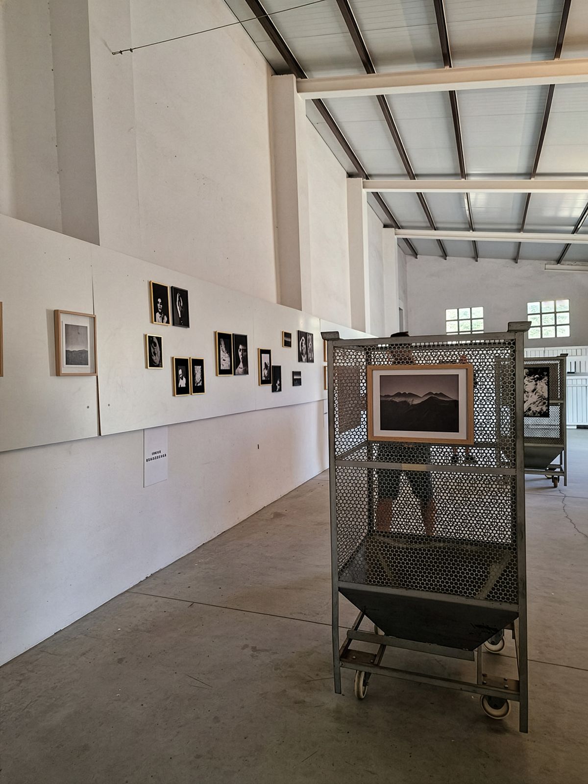 Interior of an art gallery with high ceilings and exposed beams. White walls display a series of framed photographs. In the foreground, a metal mesh display stand on wheels holds a framed landscape photograph. The concrete floor and industrial-style windows suggest a converted warehouse space.