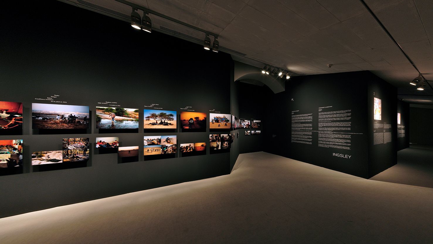 A gallery showcasing a series of photographs and descriptive text panels on dark walls, illuminated by focused spotlights.