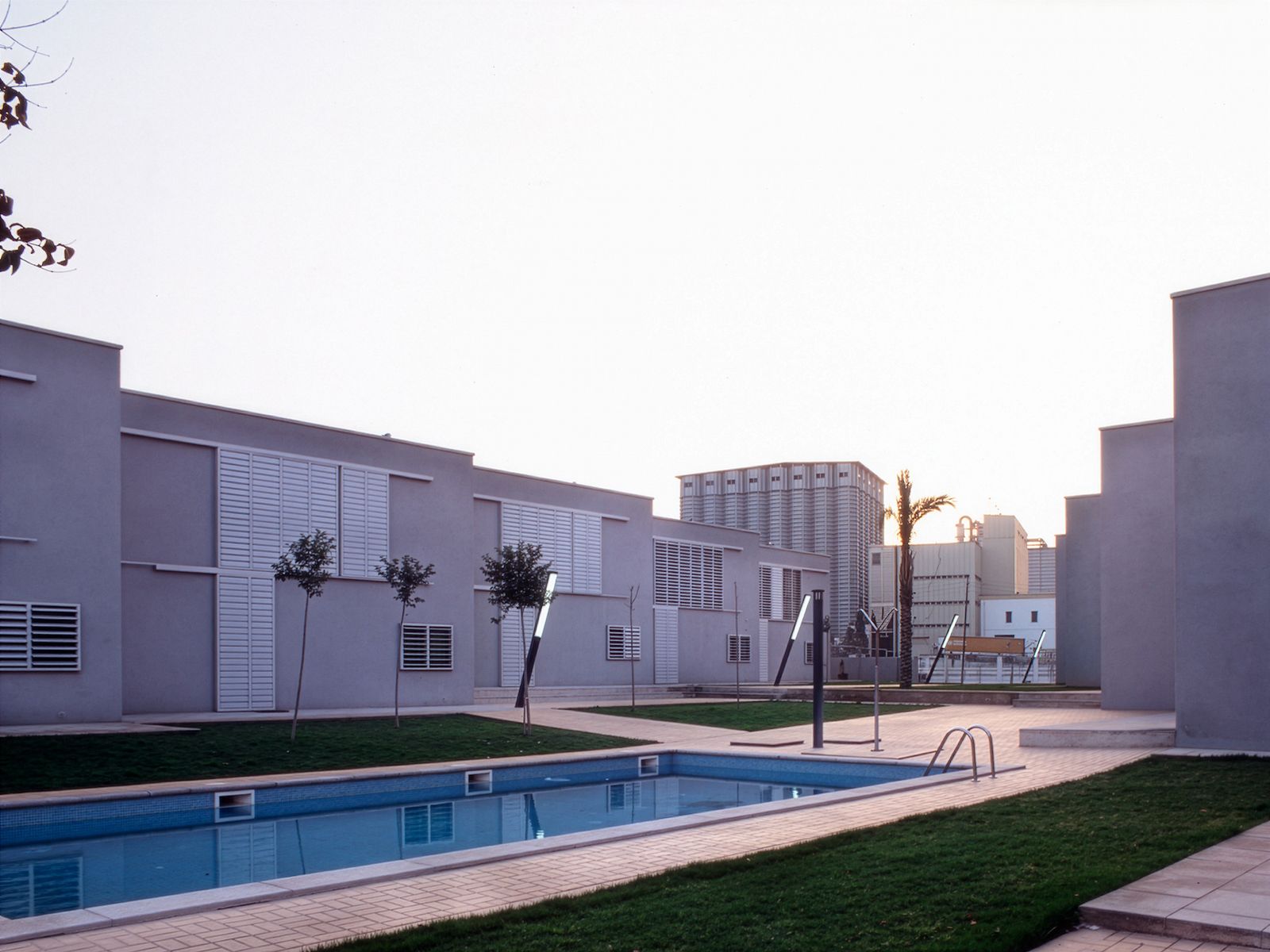 Complejo residencial moderno con edificios de techo plano, ventanas con lamas, una piscina central y áreas de césped ajardinadas.