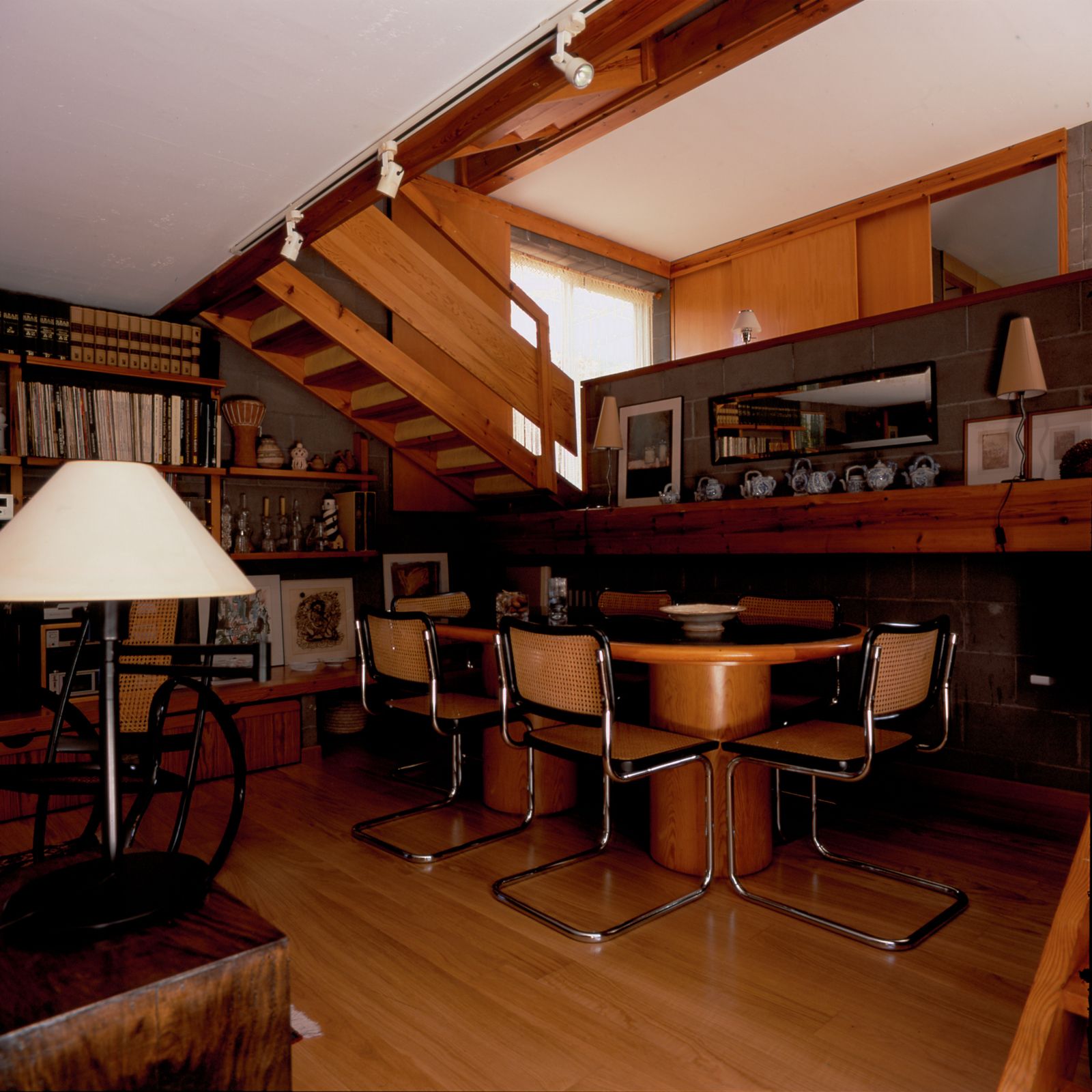Dining room with an oval wooden table, six Cesca chairs, wooden staircase leading to the upper floor, bookshelves, and various decorative items on the shelves.