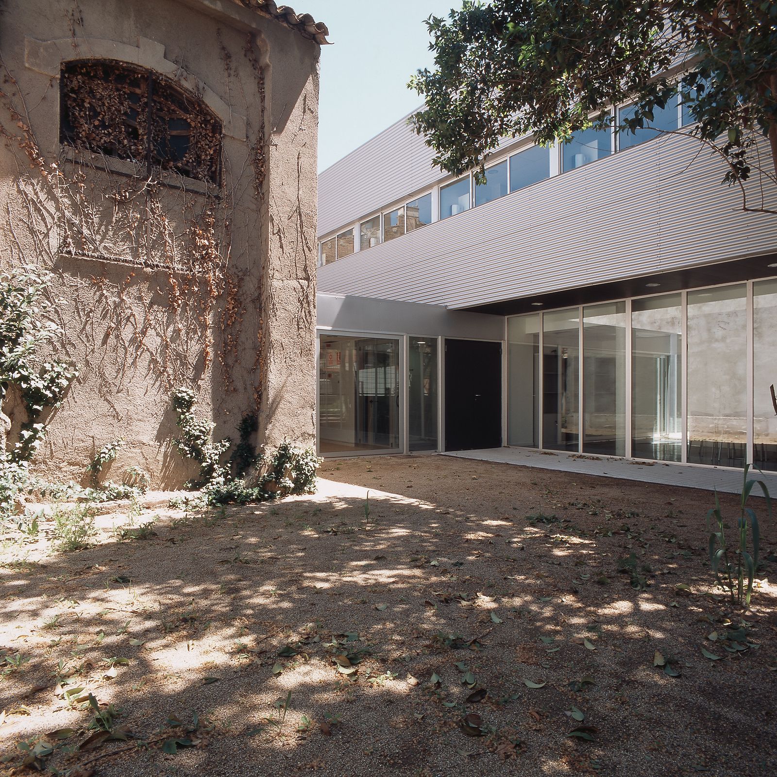 Un patio que combina arquitectura antigua y nueva, con un edificio histórico a la izquierda y una estructura moderna de vidrio a la derecha, rodeado de vegetación y árboles que dan sombra.